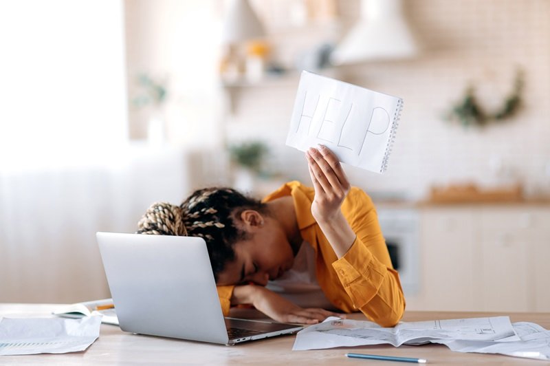 A person in a yellow shirt appears overwhelmed while working at a home office desk, resting their forehead on one hand and raising the other hand, which is holding up a piece of paper with the word 'HELP' written on it. Surrounding the laptop are various papers and documents, suggesting a state of stress or a need for assistance.